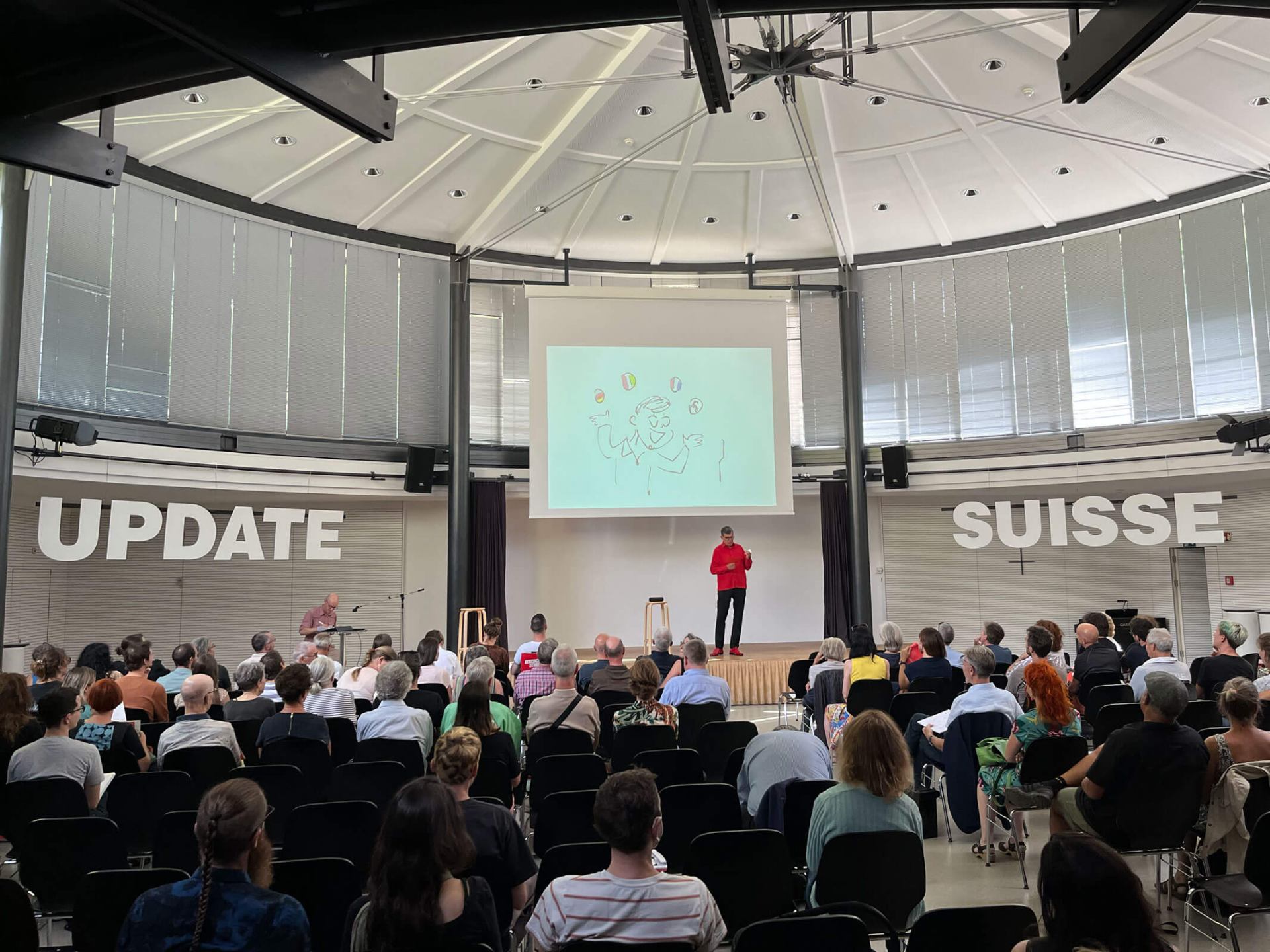 Person auf der Bühne und Publikum davor am Kick-off in Bern. Seitlich der Bühne stehen in sehr grossen Buchstaben die Worte «Update» und «Suisse».